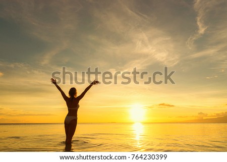 Back of young woman standing in water with raised arms and looking at sea and sunset.