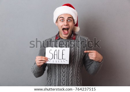 Picture of young surprised excited man wearing christmas santa hat standing isolated over grey wall holding paper with sale text. Looking camera pointing to text.