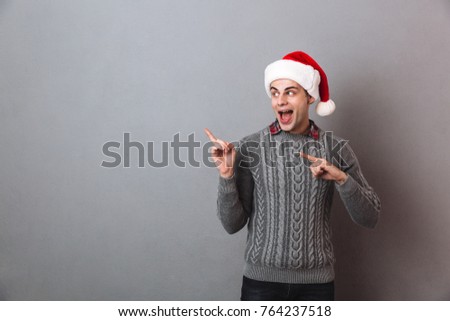 Image of young excited happy emotional man wearing christmas santa hat standing isolated over grey wall. Looking aside pointing to copyspace.