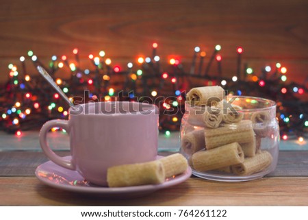 Festive still life with a pink cup of tea, a sauceer and a teaspoon, bowl of fresh crispy cookies and fairy lights, colored wooden background