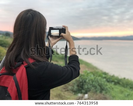Young woman backpacker with black t shirt and red bag holding camera in her hand taking a nice picture of colorful sky, river and mountain along the shore