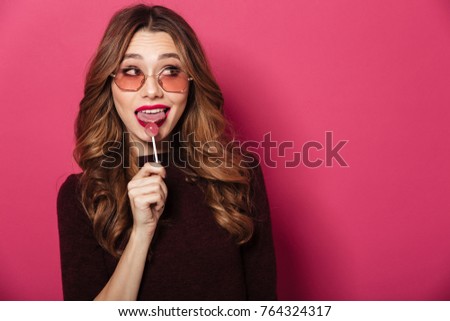 Photo of young beautiful lady wearing glasses standing isolated over pink background. Looking aside eating candy.