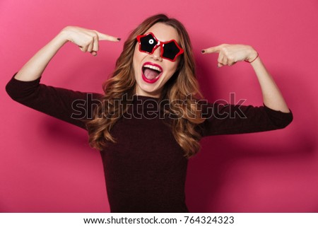 Photo of young smiling laughing lady wearing sunglasses standing isolated over pink background. Looking camera pointing to glasses.