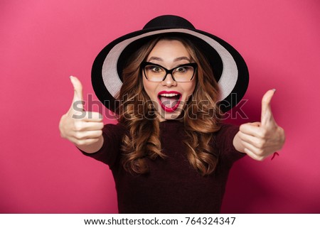 Image of young happy lady wearing hat and glasses standing isolated over pink background. Looking camera showing thumbs up.