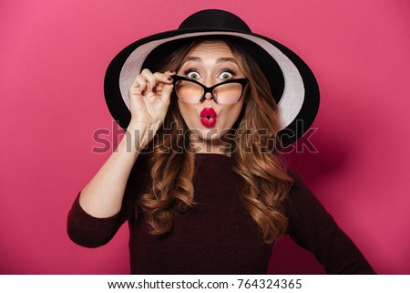 Picture of young shocked lady wearing hat and glasses standing isolated over pink background. Looking camera.