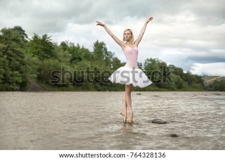 Charming ballerina stands on the pointes in the shallow river on the background of green shore and cloudy sky. She wears white tutu, pink leotard and beige ballet shoes. Her arms outstretched upward.