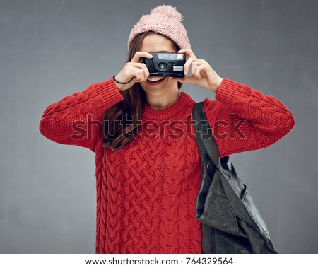 Happy traveler woman takes pictures with camera. Girl wearing warm clothes. Portrait against gray wall.