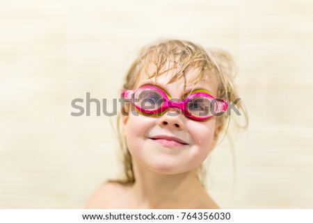 Little cute caucasian girl with blond wet hair wearing pink swimming goggles having fun and peaking out of bath side with tricky sight.