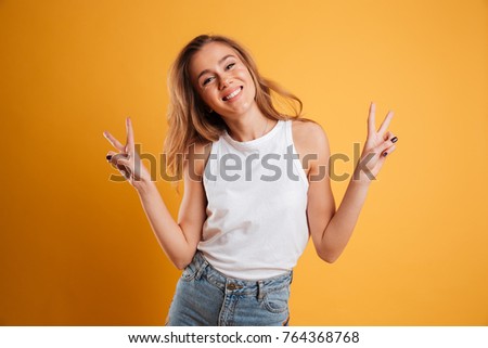 Portrait of a happy young girl showing peace gesture and looking at camera isolated over yellow background Royalty-Free Stock Photo #764368768