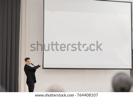 speaker conducts the business of the conference standing in front of a large white screen on the stage in the conference room
