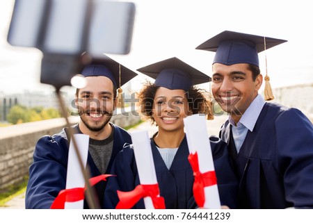 education, graduation, technology and people concept - group of happy international students in mortar boards and bachelor gowns with diplomas taking picture by smartphone selfie stick outdoors
