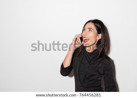 Picture of young emotional woman with red lips standing isolated over white wall. Looking aside talking by mobile phone.