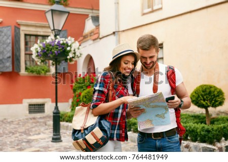 Beautiful Tourist Couple Traveling Using Map And Phone. Portrait Of Smiling Man And Young Woman Standing On Street, Searching Destination Location On Maps. Travel Concept. High Quality Image.