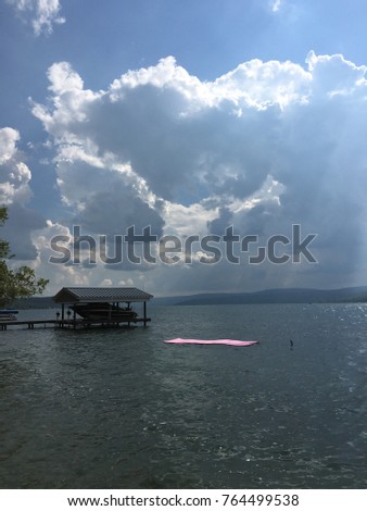 A Lone pink float is in the lake near a boathouse as the skies threaten the lake with darkening clouds, rays of sunshine and rain.