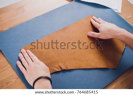 Working day in studio. Man leather worker drawing sketch of purse in notebook on table with leather wallets and tools for sewing. Effective work in leather workshop. Design of fashion leather purse