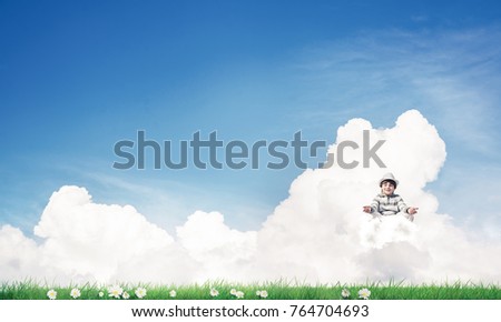Young little boy keeping eyes closed and looking concentrated while meditating on cloud in the air with bright and beautiful landscape on background.