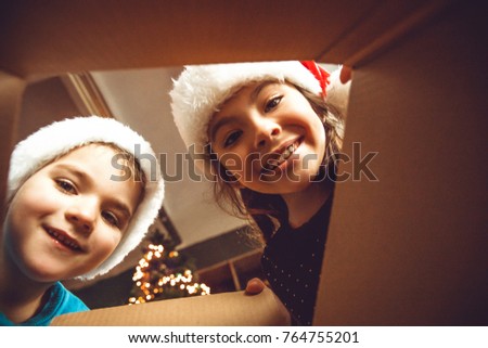 Surprised cute children girl and boy opening a Christmas present. Little kid having fun near decorated tree indoors.  Happy  holidays and New Year.