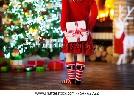 Child at Christmas tree and fireplace on Xmas eve. Little girl holding present box. Child with gift. Family with kids celebrating Christmas at home. Gifts for winter holidays at fire place. Back view.