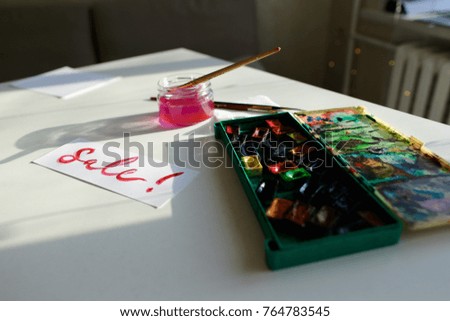Bright inscription for sale sale, written in pink watercolor paints and brush, which lies on white table in art studio. Concept of fine arts and bright colors, beautiful writing, calligraphy and