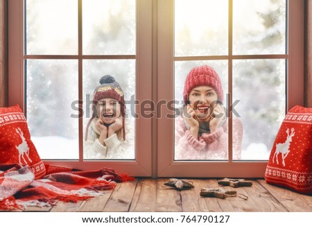 Happy loving family! Mother and child girl having fun, playing and laughing on snowy winter walk in nature. Frost season. Child and mom looking in windows, standing outdoors. View indoors home.