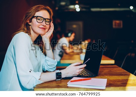 Portrait of smiling young woman in eyewear looking at camera waiting for multimedia synchronization using modern technologies,positive female freelancer share files connecting mobile to netbook
