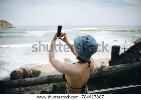 Young woman taking a photo on cellphone of the Atlantic ocean in Asturias, Spain. Travelling girl standing on hill and taking photo of the landscape
