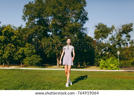 Young pretty girl with long brown hair dressed in light clothes staying on green lawn grass in the park on trees background. Summer sunny time