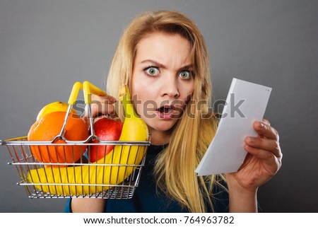 Shocked woman holding shopping basket with fruits looking at bill receipt being scared of huge prices