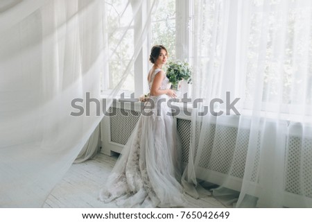 A beautiful bride in a beautiful wedding dress with lace stands by the window. White interior.