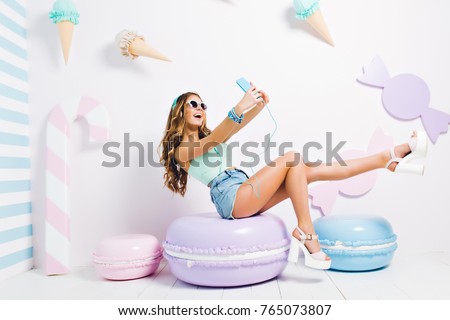 Inspired funny girl wearing tank-top and denim shorts sitting on toy macaroon and making selfie. Laughing young lady in sunglasses and headphones taking picture of herself in room with sweet interior.