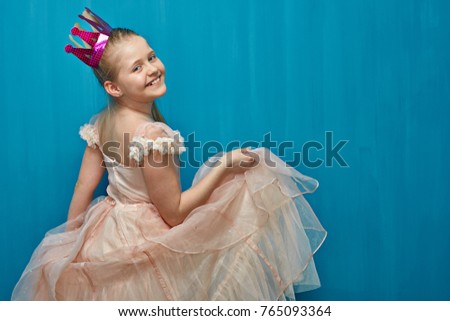 Smiling girl wearing pink dress dancing against blue wall background. Little princess with paper crown.
