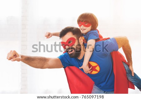 Father and son in the red and blue suits of superheroes. On their faces are masks and they are in raincoats. They are posing in a bright room. Royalty-Free Stock Photo #765097198
