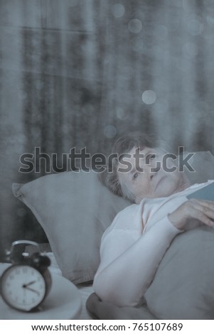 Photo through window of clock on nightstand next to sleepy grandmother in bed