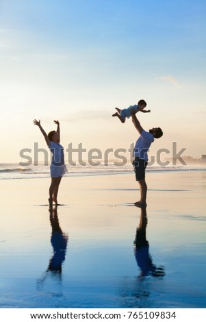Father tossing high in air baby son, mother jumping by water pool. Happy family walk with fun by sunset black sand beach with sea surf. Active parents, outdoor activity on summer vacation with kids.
