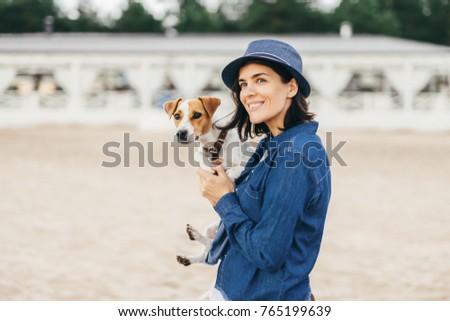 Beautiful woman wears fashionable hat and denim shirt, has walk with her dog outdoor, find out unknown places, looks thoughtfully into distance, has happy expression and pleasant smile