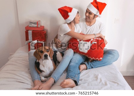 young couple sitting on bed with christmas santa hats having fun with their dog. Presents on bed. Home, indoors. Xmas concept