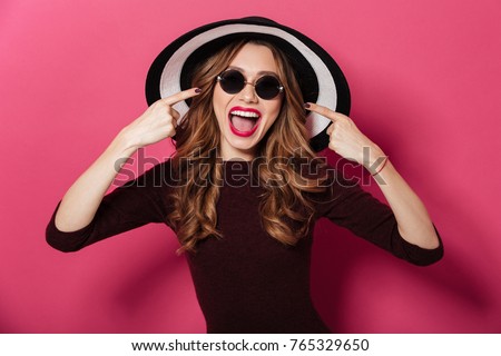 Picture of young happy lady wearing hat and sunglasses standing isolated over pink background. Looking camera pointing.