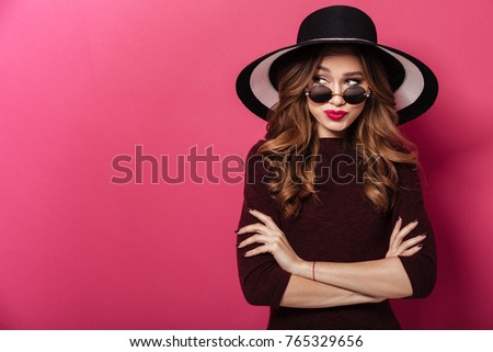 Photo of young confused lady wearing hat and sunglasses standing isolated over pink background. Looking aside.