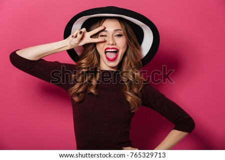 Image of young pretty lady wearing hat standing isolated over pink background. Looking camera showing peace gesture.