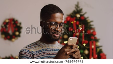 Close up of young African American man in glasses taping on his smart phone and smiling. Christmas tree on the background. Indoor. Portrait