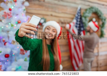 A young girl in a santa hat makes a selfie on the background of a man who hangs an American flag on the wall.