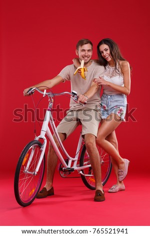 Young couple on a red background, on a bicycle with a banana, looking at the camera ..