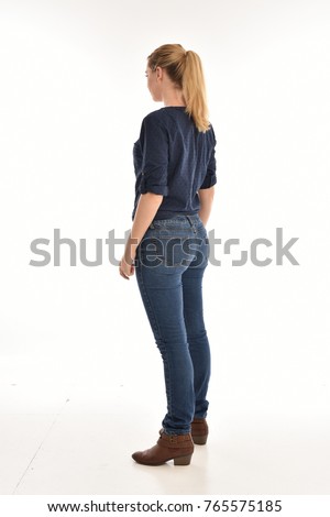 Full length portrait of a girl wearing simple blue shirt and jeans, standing pose facing away on a white background.
 Royalty-Free Stock Photo #765575185