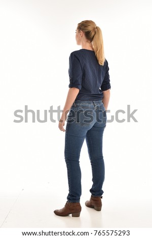 Full length portrait of a girl wearing simple blue shirt and jeans, standing pose facing away on a white background.
 Royalty-Free Stock Photo #765575293
