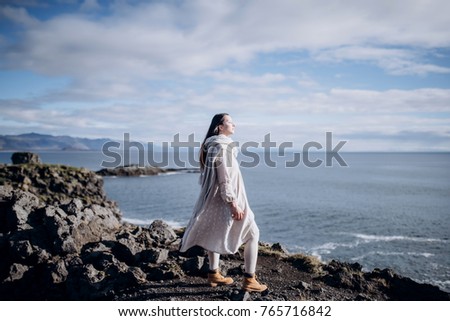 Brunette long hair woman in an amazing linen dress and beige woolly scarf is standing on a cliff and looking to the ocean. Sunny day, blue cloudy sky. Dream travel concept. film photo Soft focus