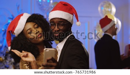 Portrait of young happy African American couple in santa clause hats taking selfies on a smart phone at the New Year or Christmas party. Dancing people background. Indoor