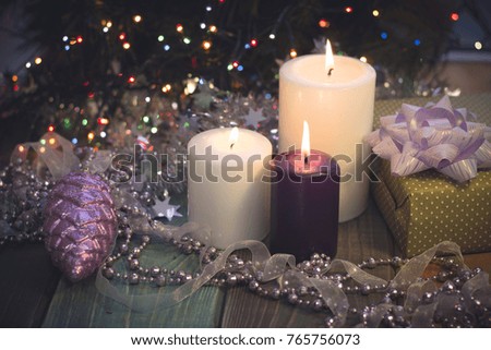 A festive still life with a purple and two white candles of different size, a handcrafted gift box with a lilac bow, a garland of a ribbon and beads and a purple pinecone. Dark Christmas background