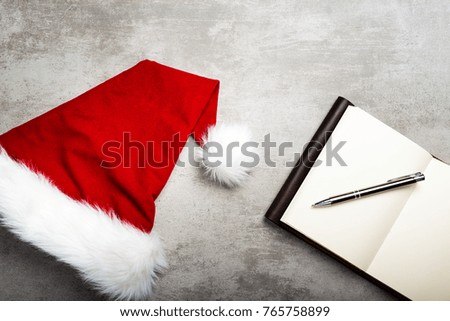 A red santa hat and a notebook with blank paper and a pen on a gray concrete table.