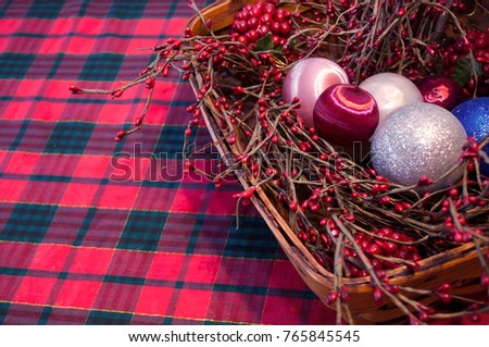 A Christmas basket filled with holly branches and tree ornaments with a plaid tablecloth background