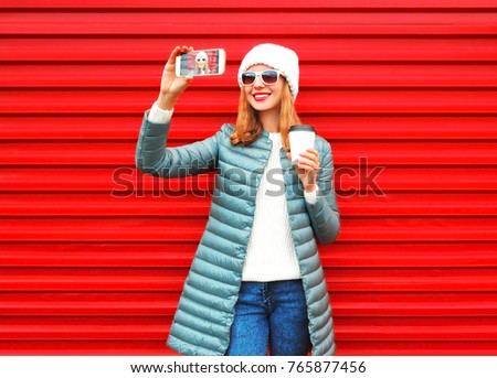 Portrait of happy smiling young woman taking selfie with smartphone wearing white hat, jacket on red background